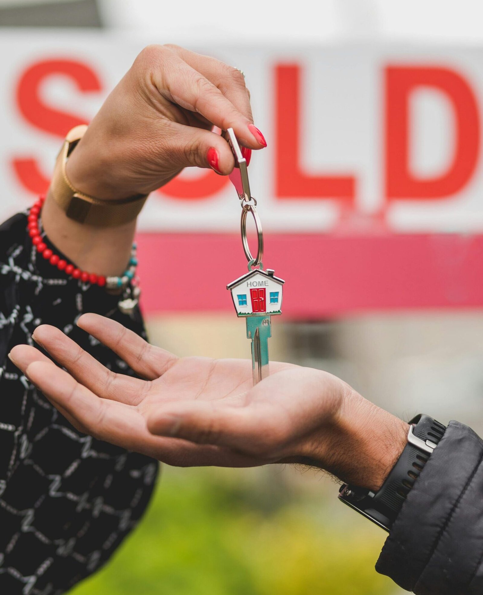 Close-up of hands exchanging house key with colorful SOLD sign in the background.